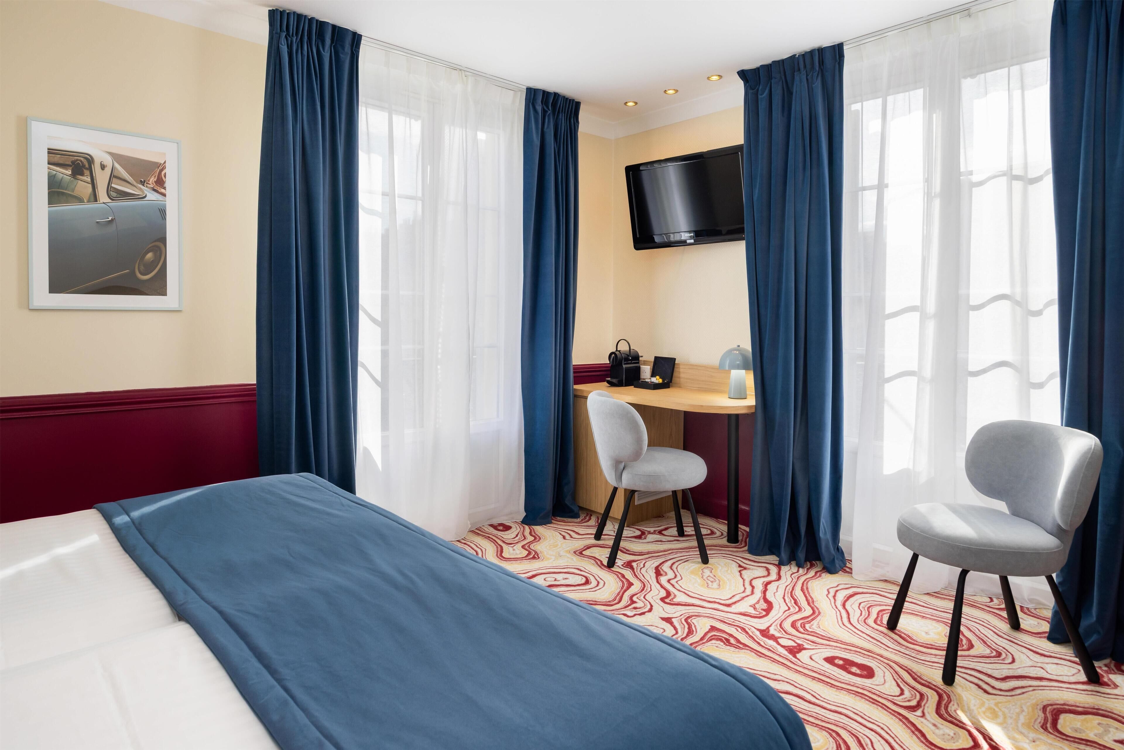 A hotel room with a bed, two chairs, a desk, a TV, and a patterned carpet.