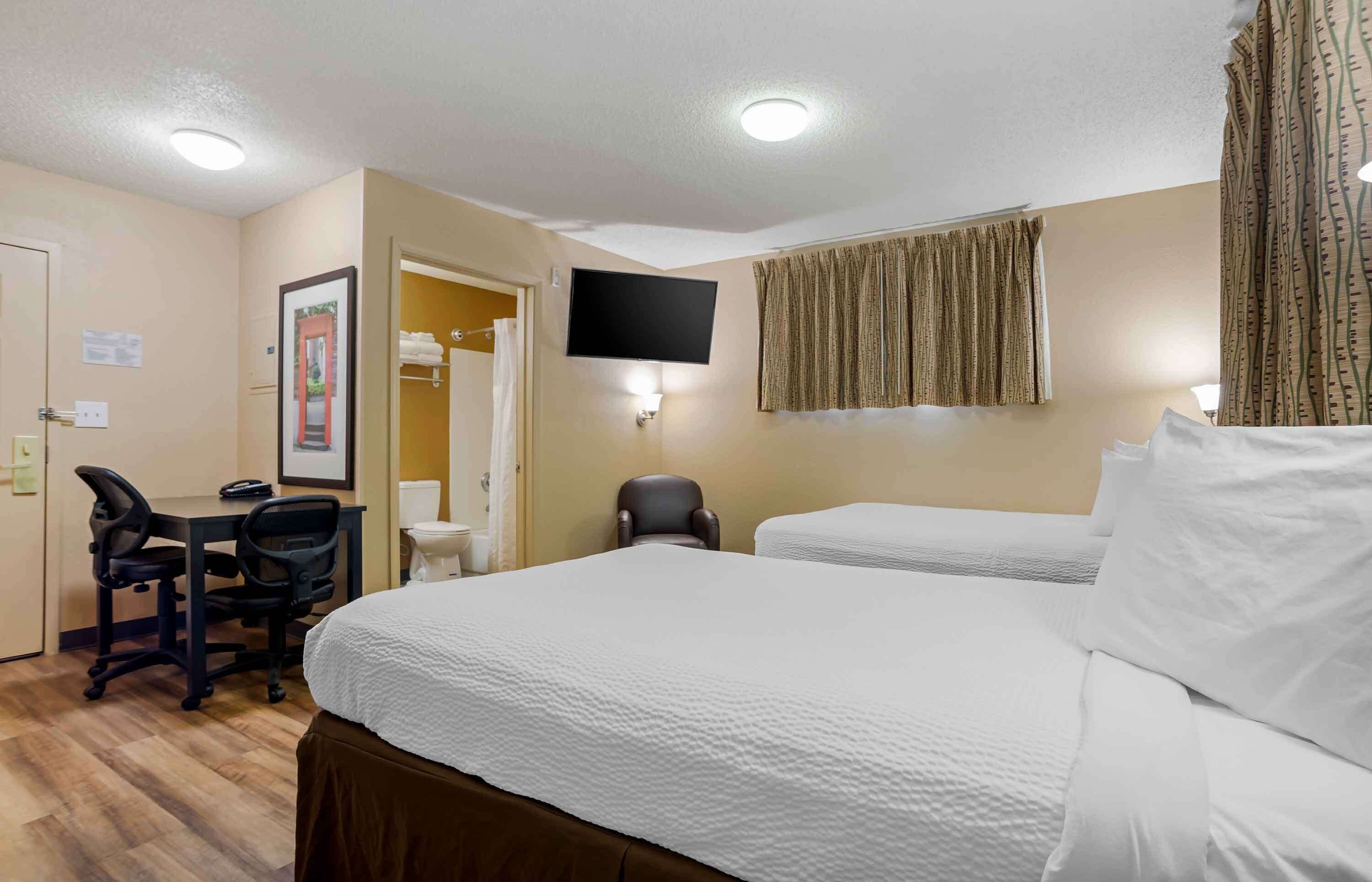 A hotel room with two beds, a desk with chairs, a TV, and a bathroom with a towel rack.