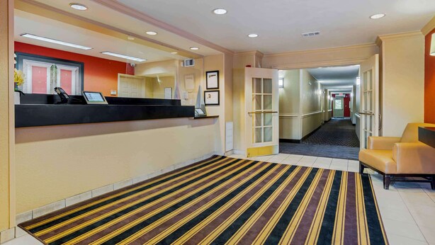 Een hotel receptie met een receptiebalie, een gestreept tapijt, een wachtruimte met een bank en een gang naar andere kamers.