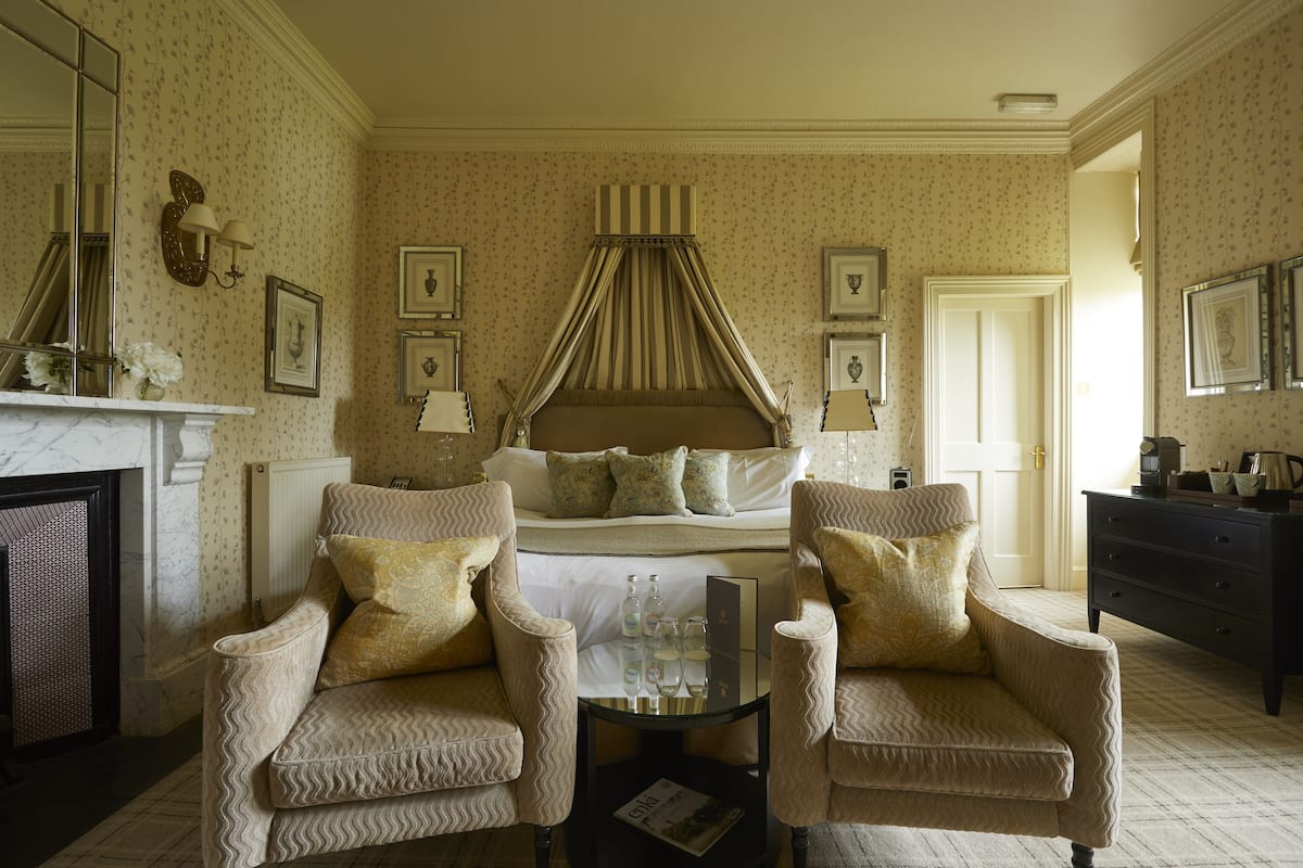 A bedroom with a four-poster bed, two armchairs, a fireplace, and a dresser.