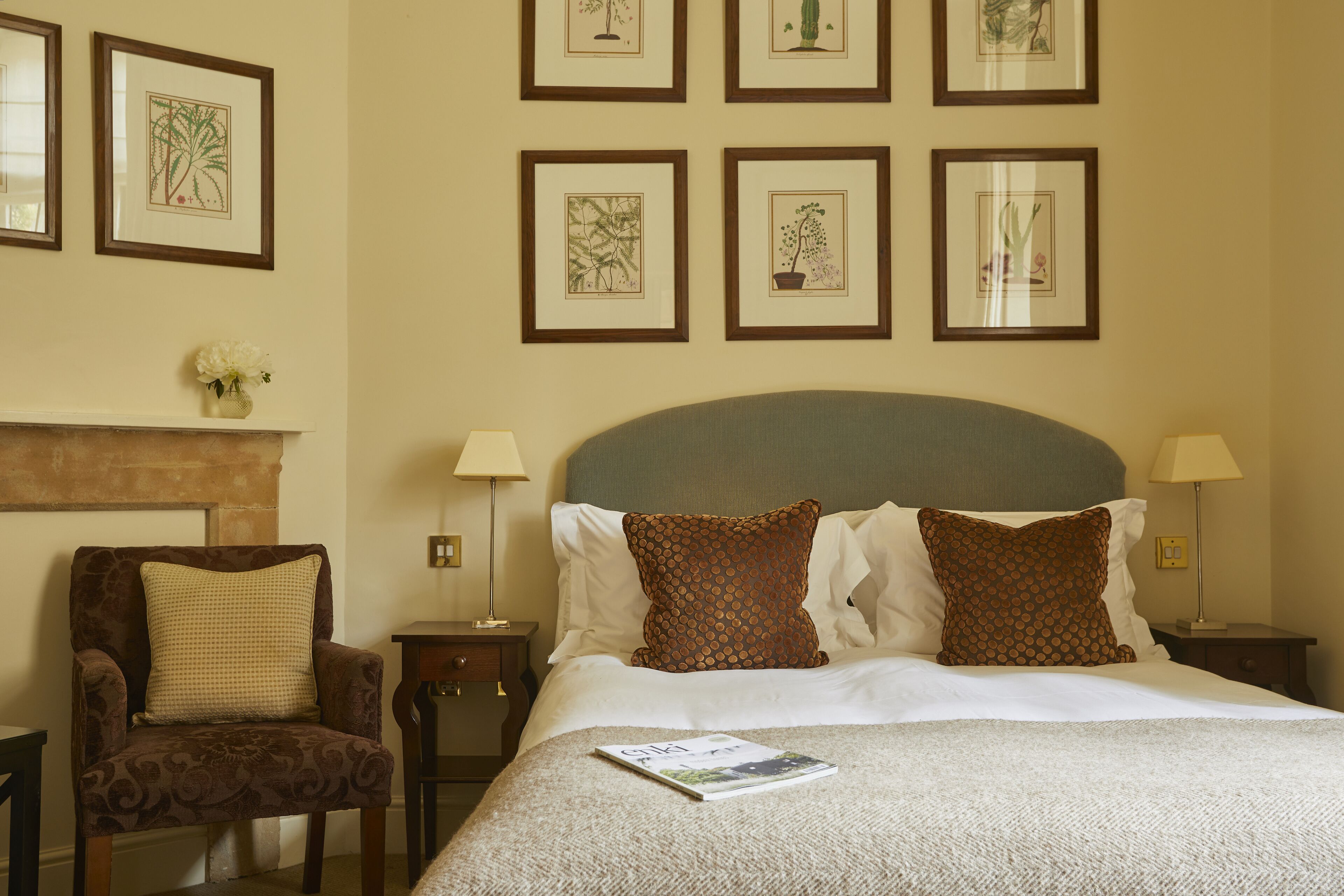 A hotel room with a large bed, two nightstands, a chair, a fireplace, and framed botanical artwork on the wall.