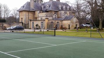 Una cancha de tenis con red, frente a un edificio de piedra grande con múltiples ventanas y una chimenea.