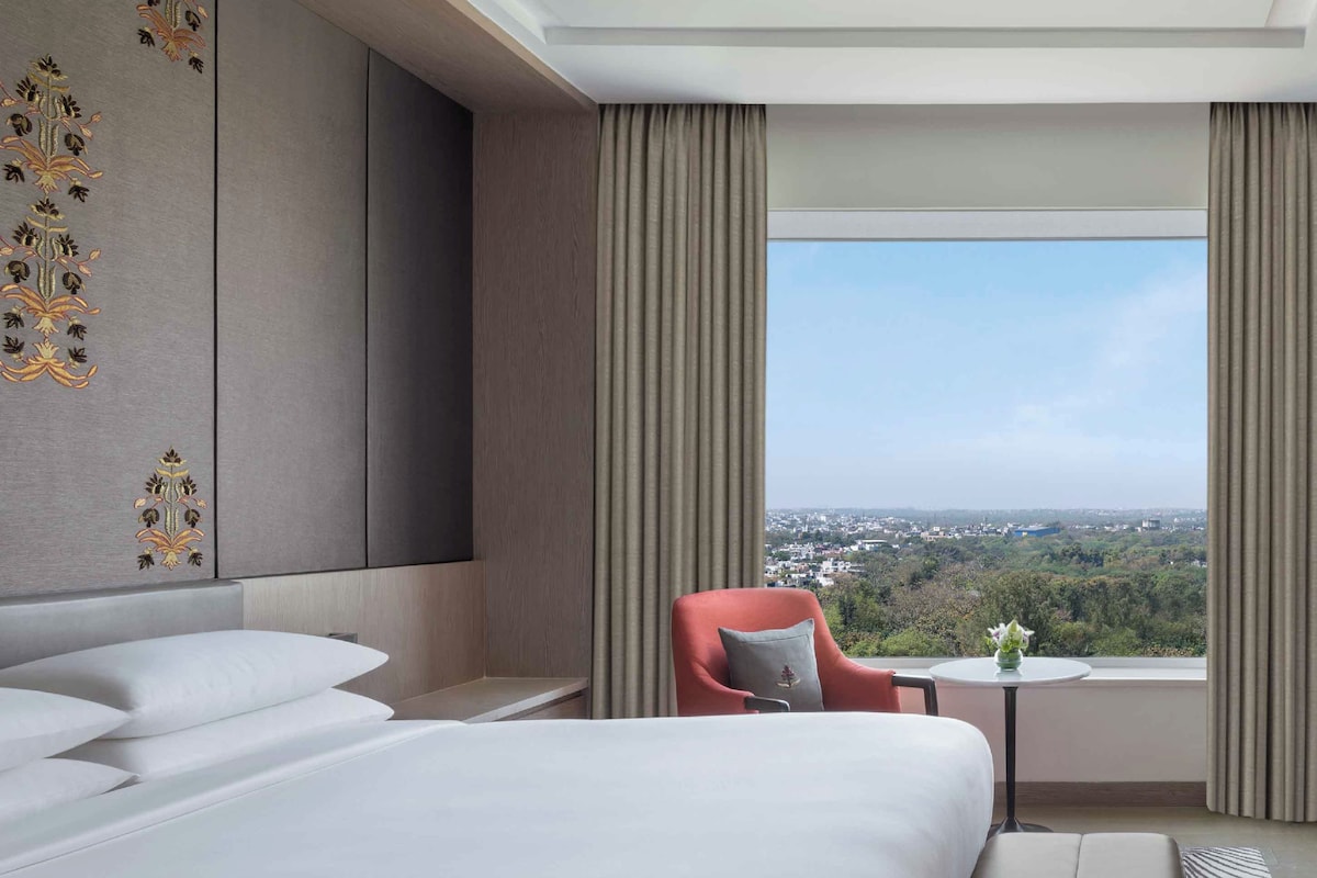 A hotel room with a large bed, a red armchair, a small table with a vase, and a view of a cityscape.
