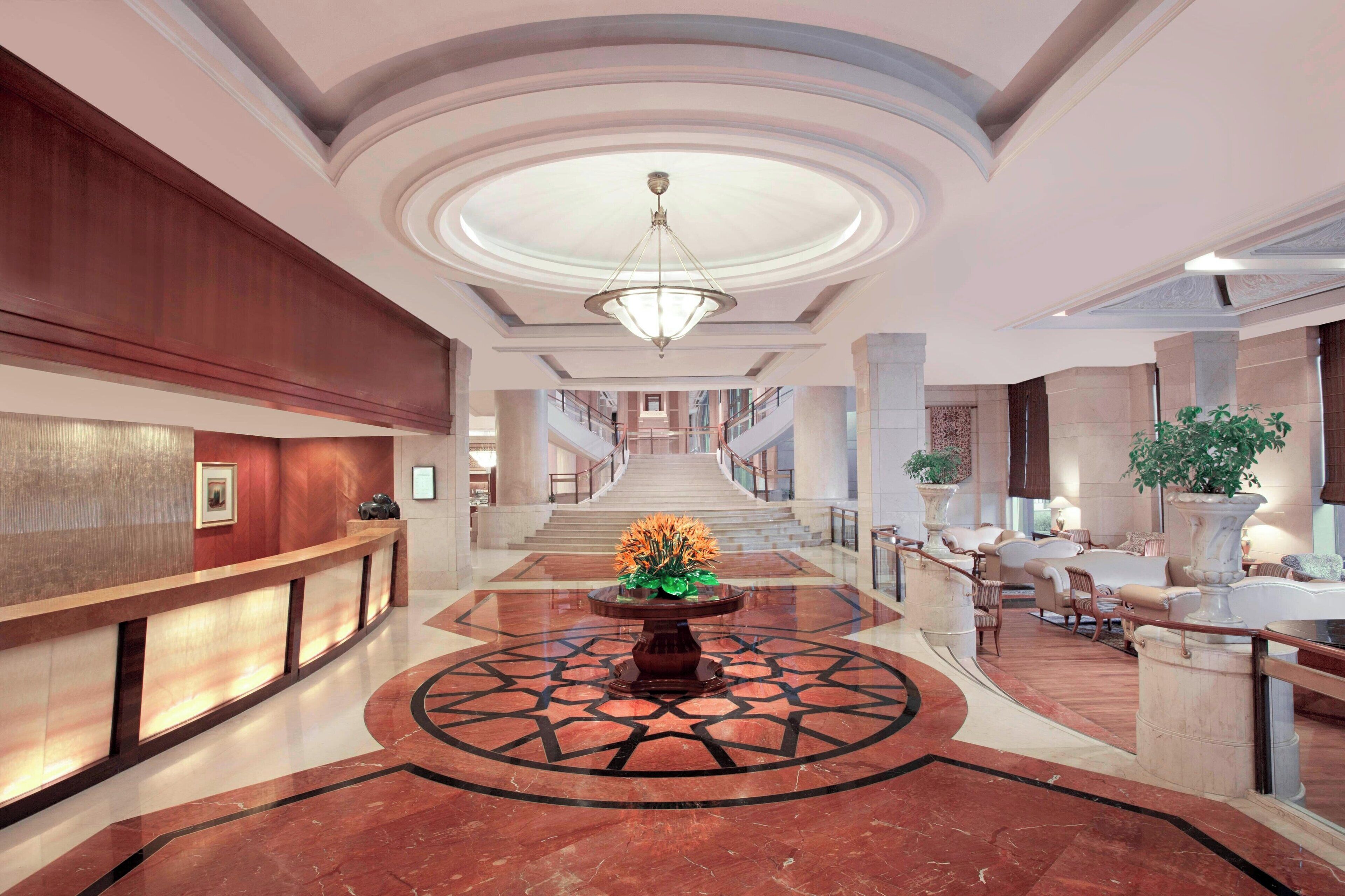 Un vestíbulo de hotel con una recepción, un arreglo floral central, una escalera y áreas de estar.