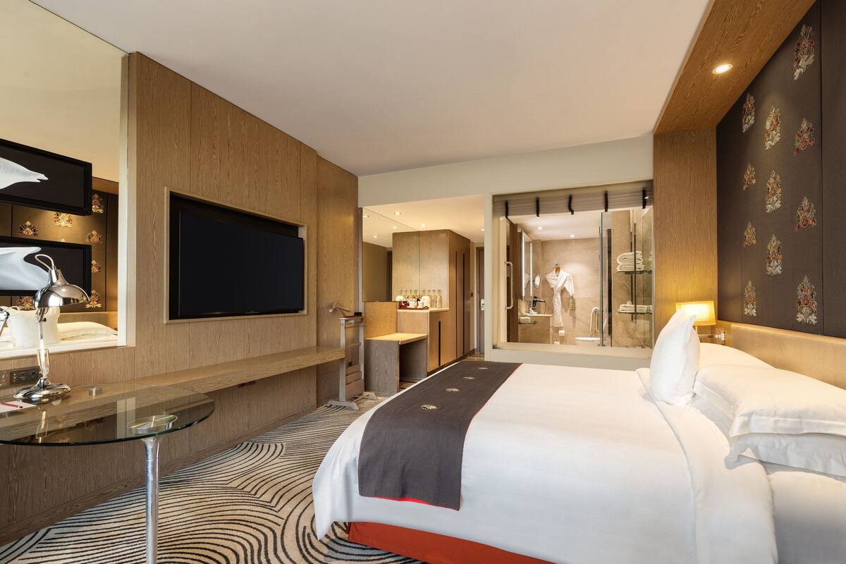 Habitación de hotel moderna con una cama grande, una mesa de vidrio, un televisor y un baño visible a través de una puerta de cristal.