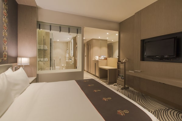 Habitación de hotel con una cama grande, un televisor montado en la pared, un baño con espejo y perchero, y una pequeña zona de estar.