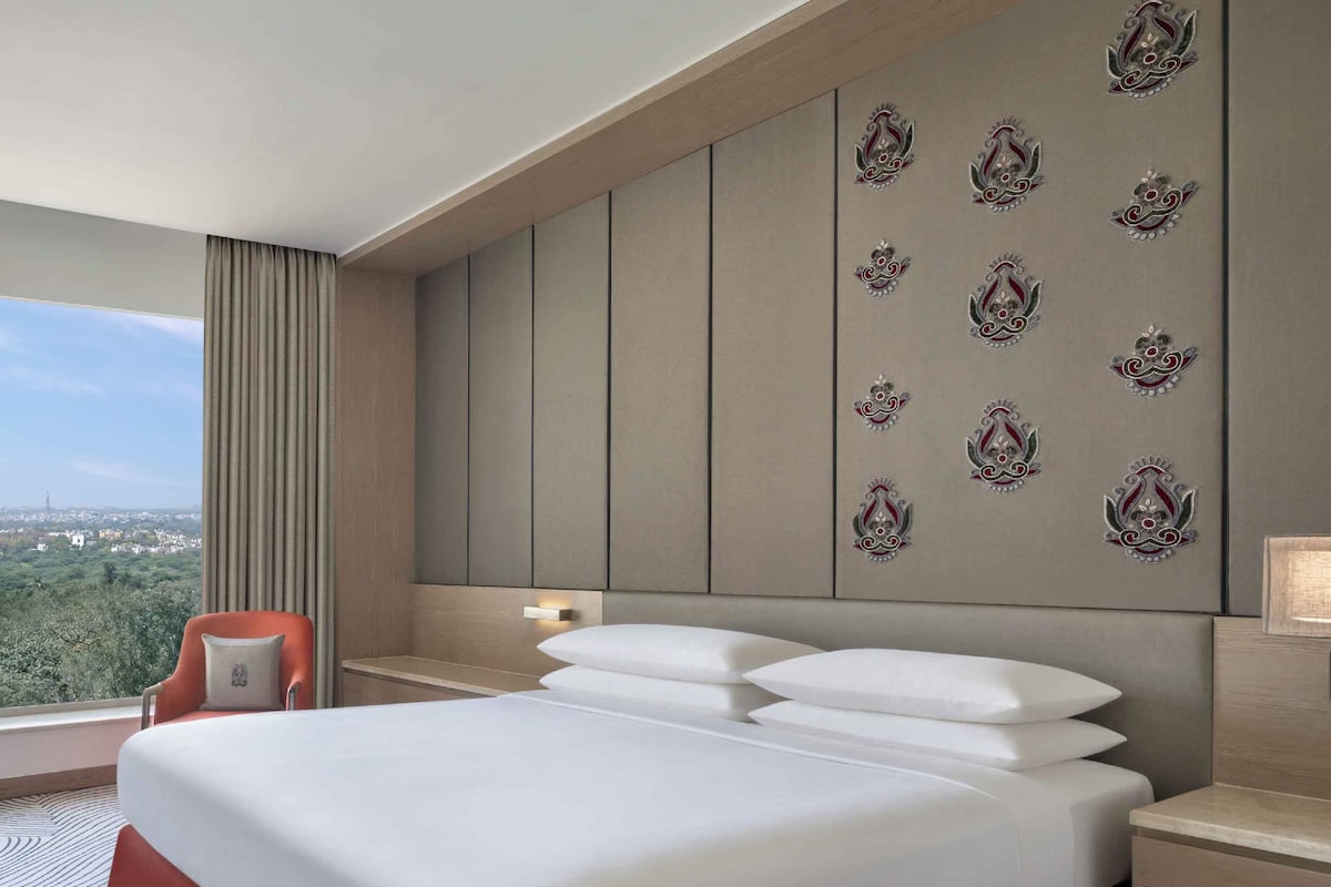 A hotel room with a large bed, a red chair, a city view, and decorative wall panels.