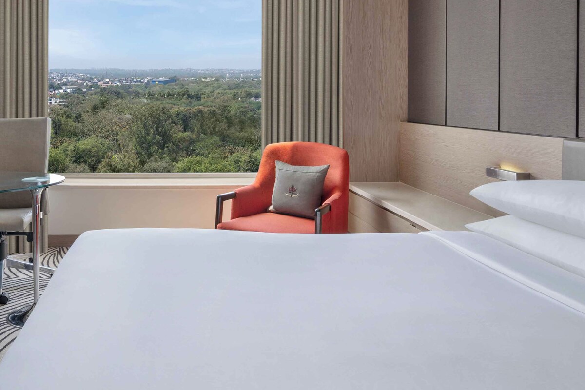 A hotel room with a large bed, a chair, a desk, and a view of a cityscape through a window.