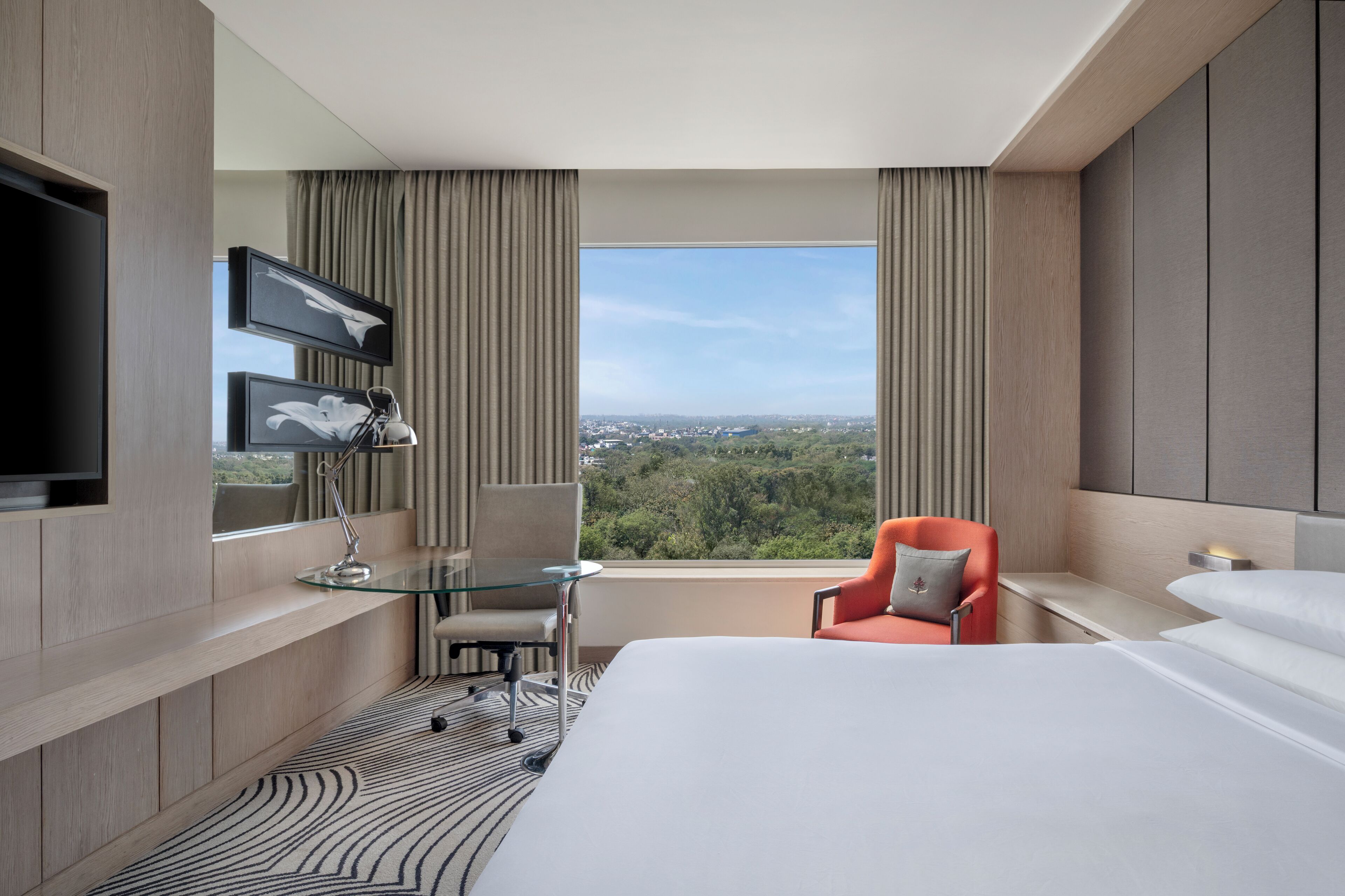 Een moderne hotelkamer met een groot bed, een glazen tafel, een stoel, een aan de muur bevestigde televisie en een uitzicht op de stad door grote ramen.