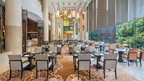 A spacious indoor area with multiple dining tables and chairs, a patterned carpet, and large windows offering a view of greenery outside. - Sheraton New Delhi Hotel (New Delhi)