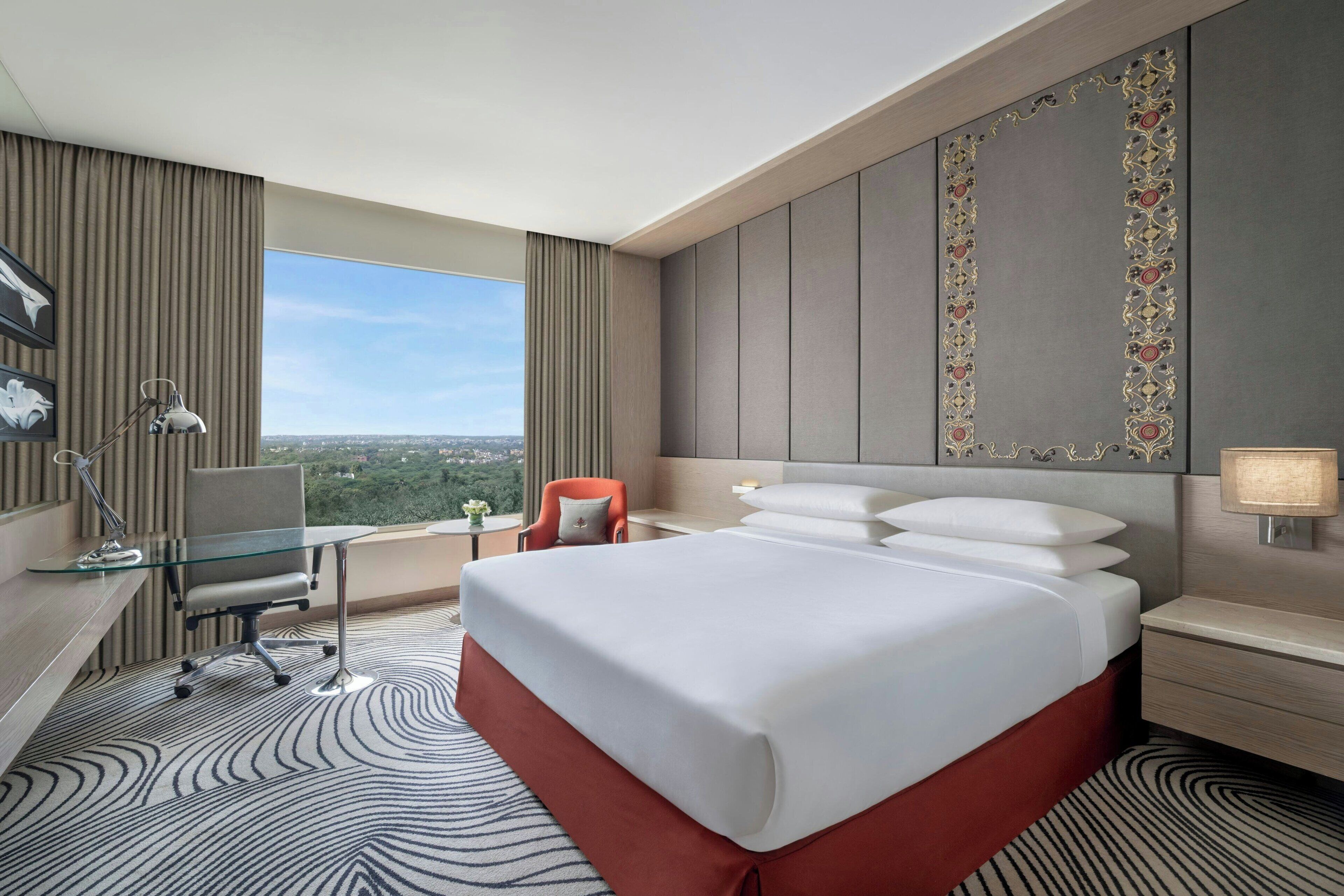 A hotel room with a large bed, a desk with a chair, a desk lamp, a nightstand, and a window with a view of the city.