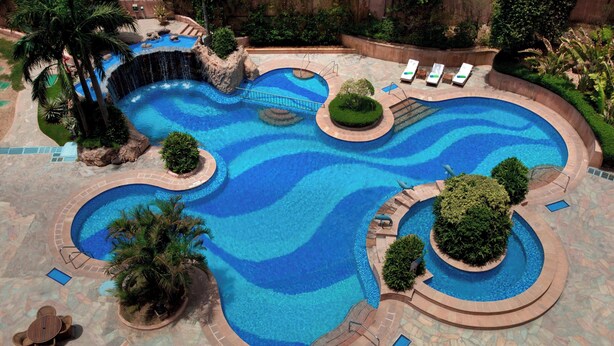A large, multi-level swimming pool with a waterfall feature, surrounded by palm trees and lounge chairs. - Sheraton New Delhi Hotel (New Delhi)