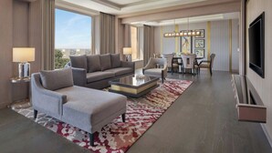 A modern hotel room with a living area featuring a sofa, armchair, and coffee table, a dining area with chairs and a table, and a large TV on the wall. - Sheraton New Delhi Hotel (New Delhi)