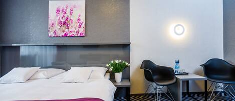 Una habitación de hotel con una cama, una mesita de noche con un jarrón de flores, dos sillas negras, una pequeña mesa con una botella y un vaso, y una lámpara de pared.