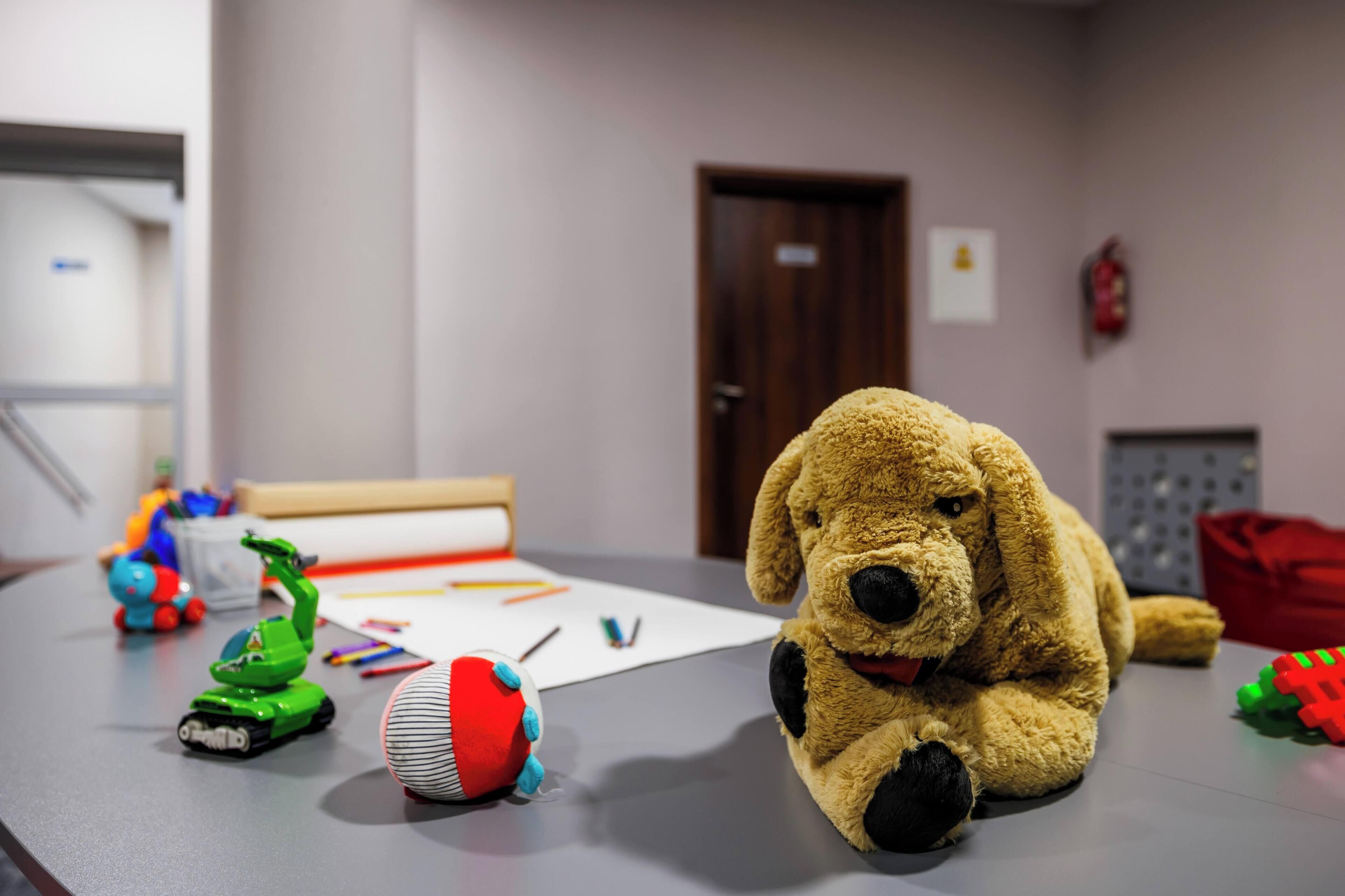 Una habitación con un perro de peluche grande, un vehículo de juguete verde y una pelota roja sobre una mesa.