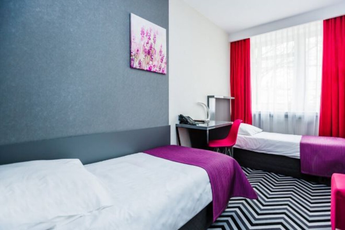 A hotel room with two beds, a desk, a chair, and a large window with red curtains.