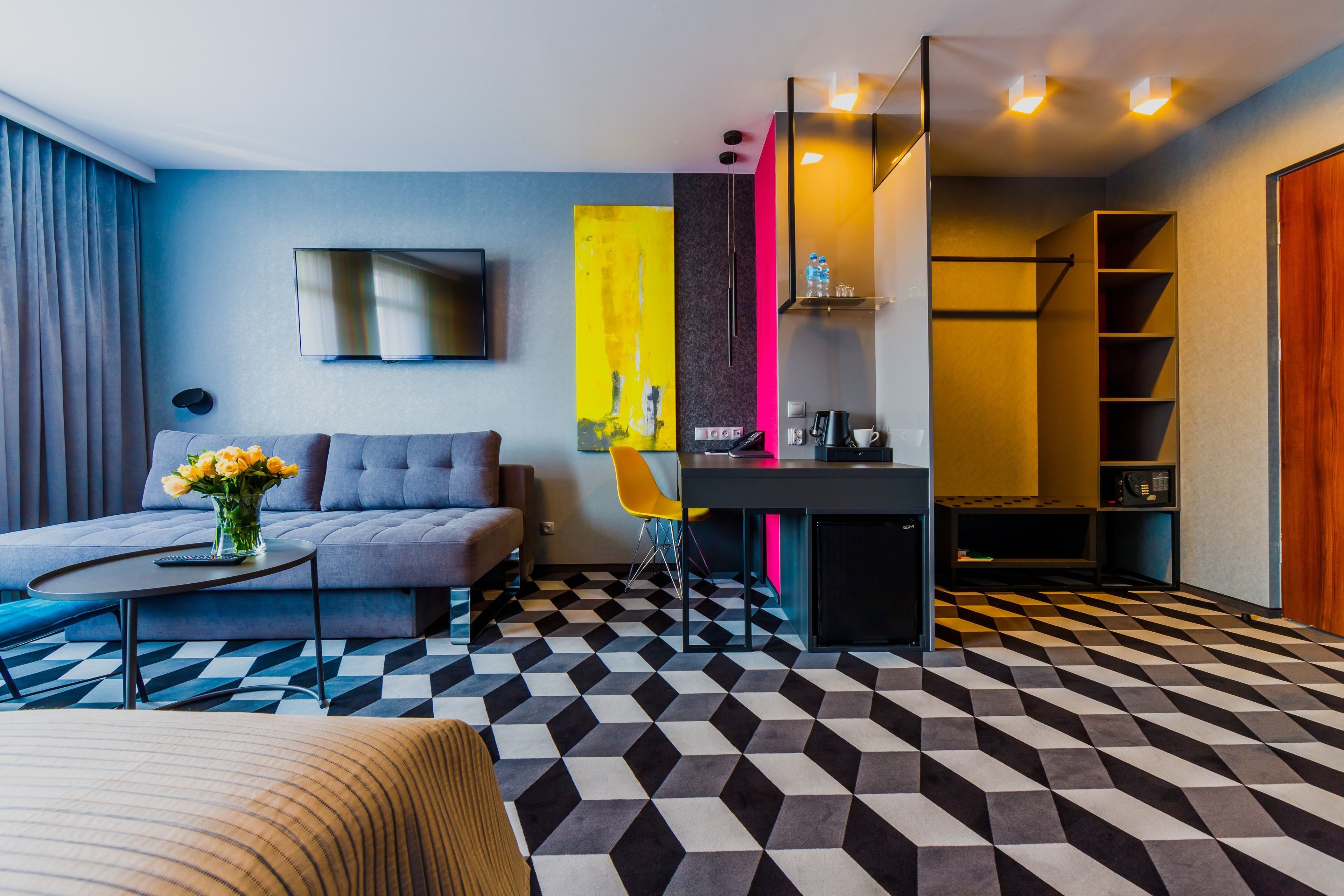 Une chambre d’hôtel moderne avec un sol à carreaux, un canapé gris, une petite table sur laquelle est posé un vase de fleurs, un téléviseur fixé au mur et un bureau avec une chaise.