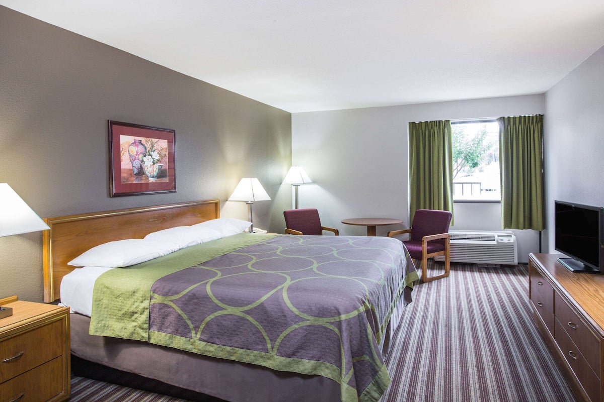 A hotel room with a bed, two chairs, a small table, a TV, and a window with curtains.