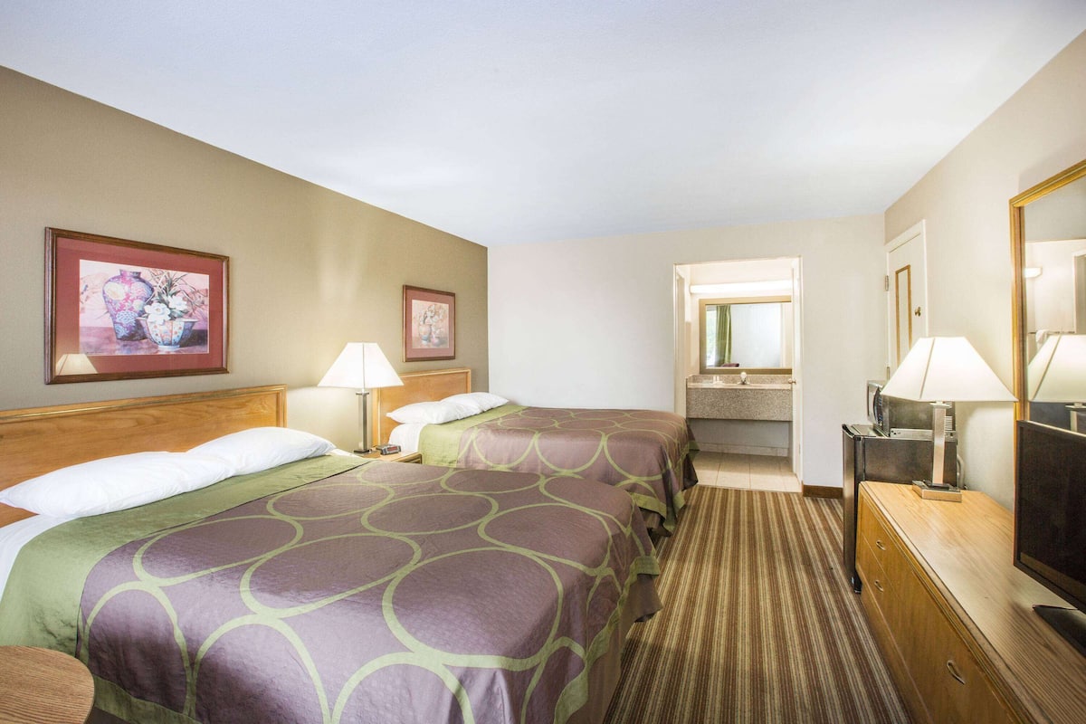 A hotel room with two beds, a wooden headboard, a bathroom with a mirror, and a television on a wooden dresser.