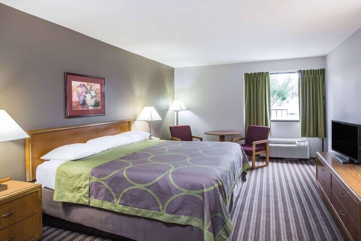 A hotel room with a bed, two chairs, a small table, a TV, and a window with curtains.