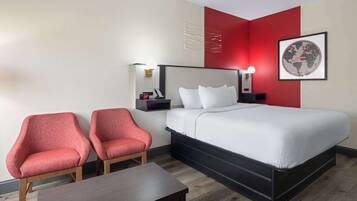 Une chambre d’hôtel avec un lit, deux fauteuils rouges, une table de chevet, une petite table avec une lampe, et une carte du monde encadrée accrochée au mur.