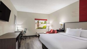 A hotel room with a large bed, a desk, a chair, a TV, and a window with a view of greenery. - Ramada by Wyndham Locust Grove (Locust Grove)