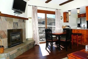 A cozy living room with a fireplace, a dining table with chairs, a TV mounted above the fireplace, and a view of the outdoors through a large window. - Rockies Condominiums by Mountain Resorts (Steamboat Springs)