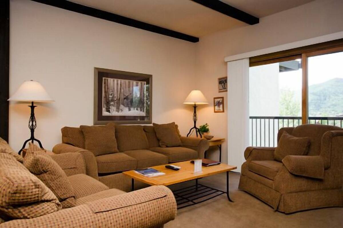 Un salon comprenant un canapé, des fauteuils, une table basse et deux lampadaires. Un tableau orne le mur et une fenêtre offre une vue sur l’extérieur.