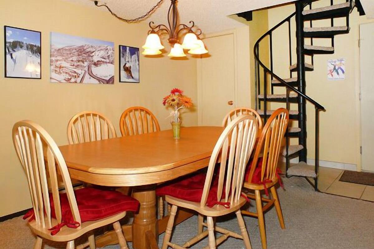 Un coin repas avec une table en bois et six chaises, un lustre et un escalier en colimaçon en arrière-plan.