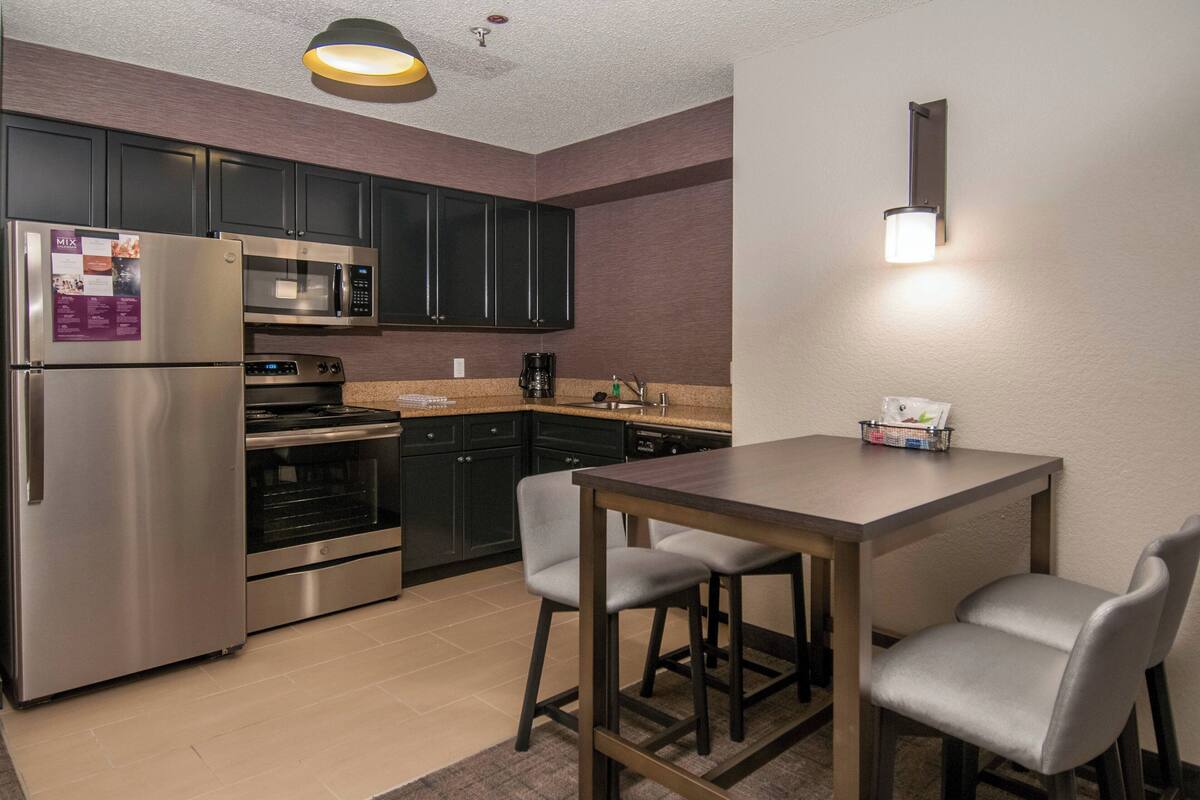 A modern kitchen with stainless steel appliances, dark cabinets, and a wooden dining table with two chairs.