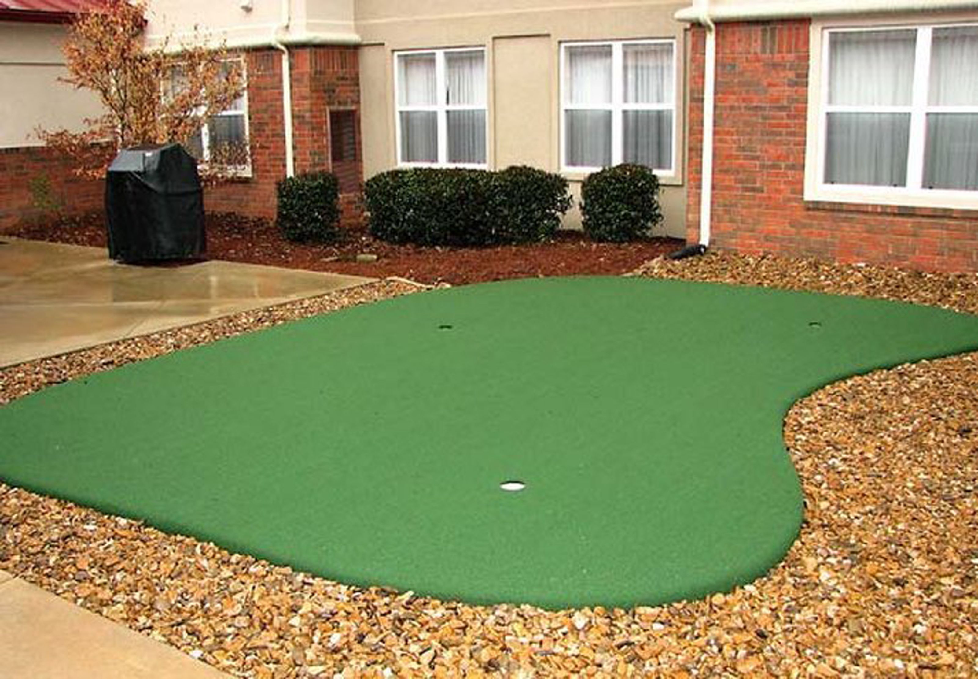 A green artificial turf putting green with a pebble border, surrounded by a brick building with windows and a hedge.