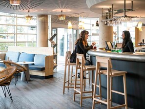 A modern bar area with wooden stools, a bar counter, and a seating area with a sofa and chairs. - Novotel Suites Paris Stade De France (Saint-Denis)