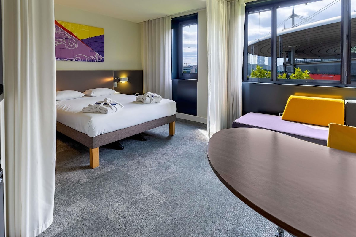 A hotel room with a bed, a table, a chair, and a view of the cityscape through large windows.