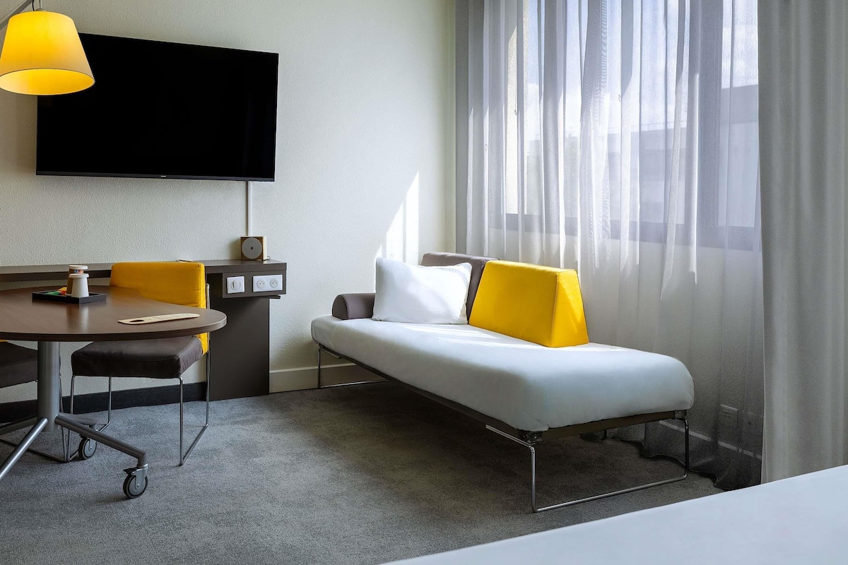 A hotel room with a bed, a desk, a chair, a television, and a window with sheer curtains.