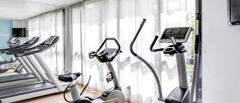 A gym with treadmills, an elliptical, and a stationary bike, with large windows and a tiled floor.