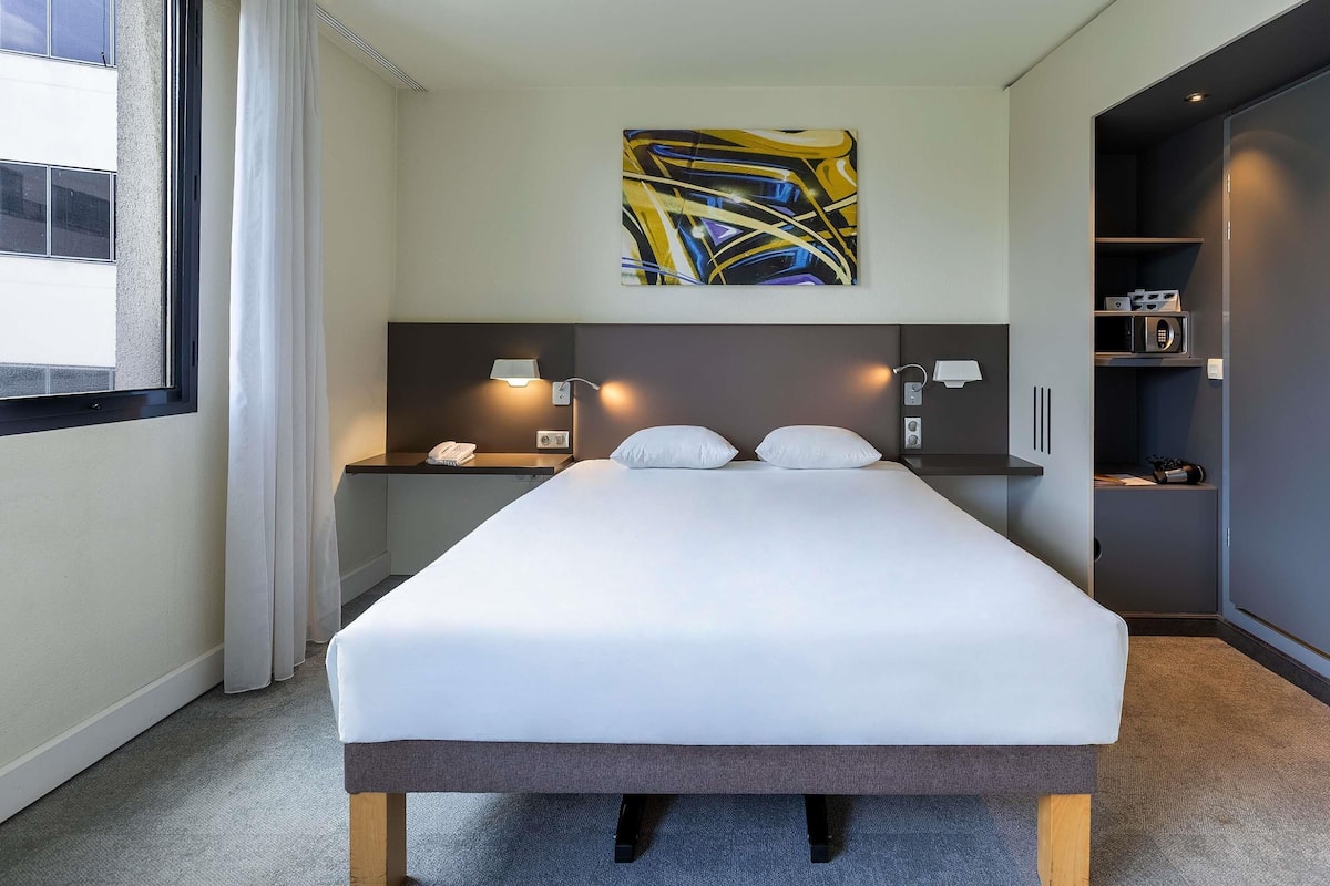 A modern hotel room with a large bed, a bedside table with a lamp, a painting on the wall, and a built-in wardrobe.
