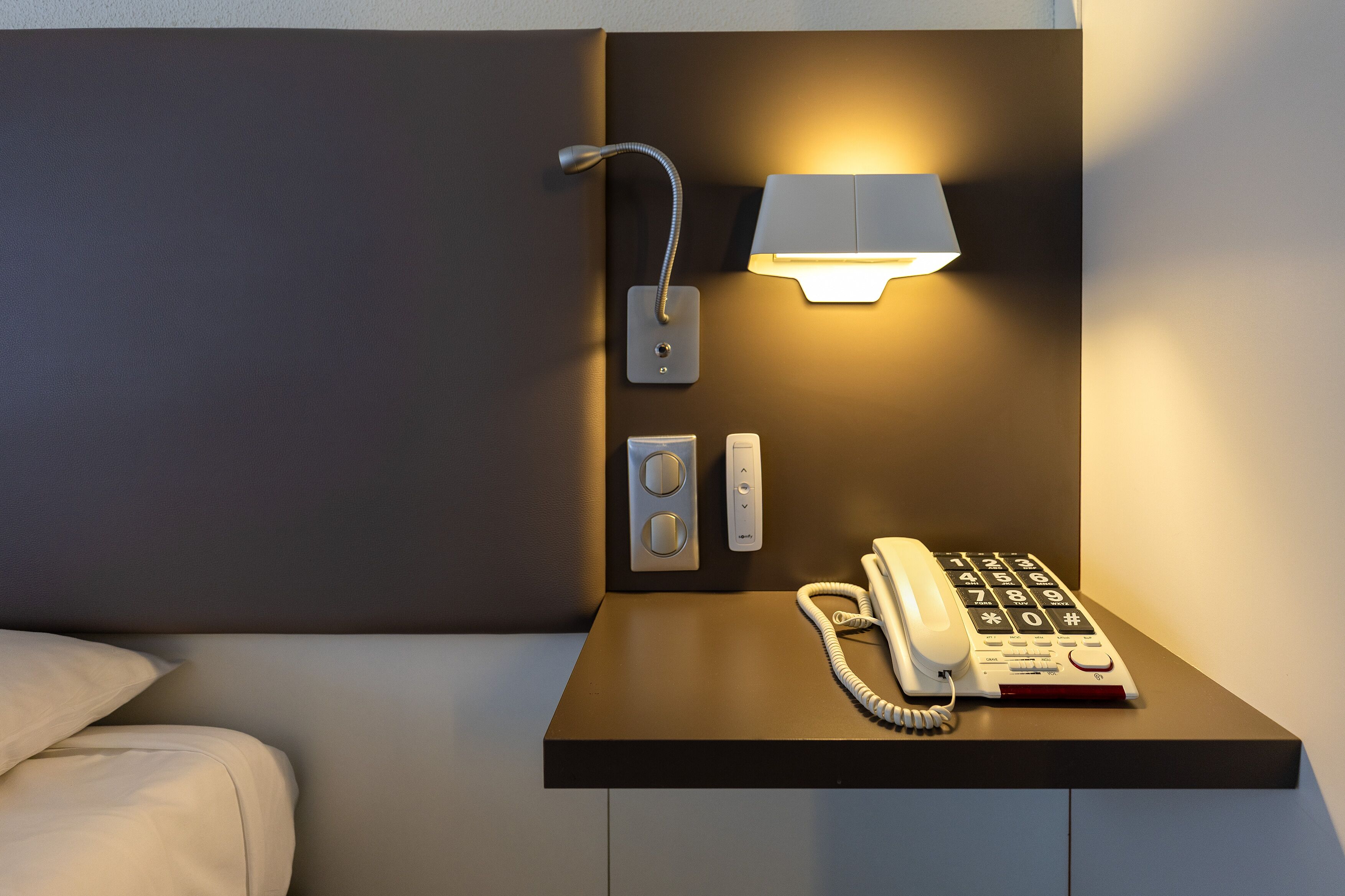 A hotel room with a bedside table, a lamp, a phone, and a control panel.