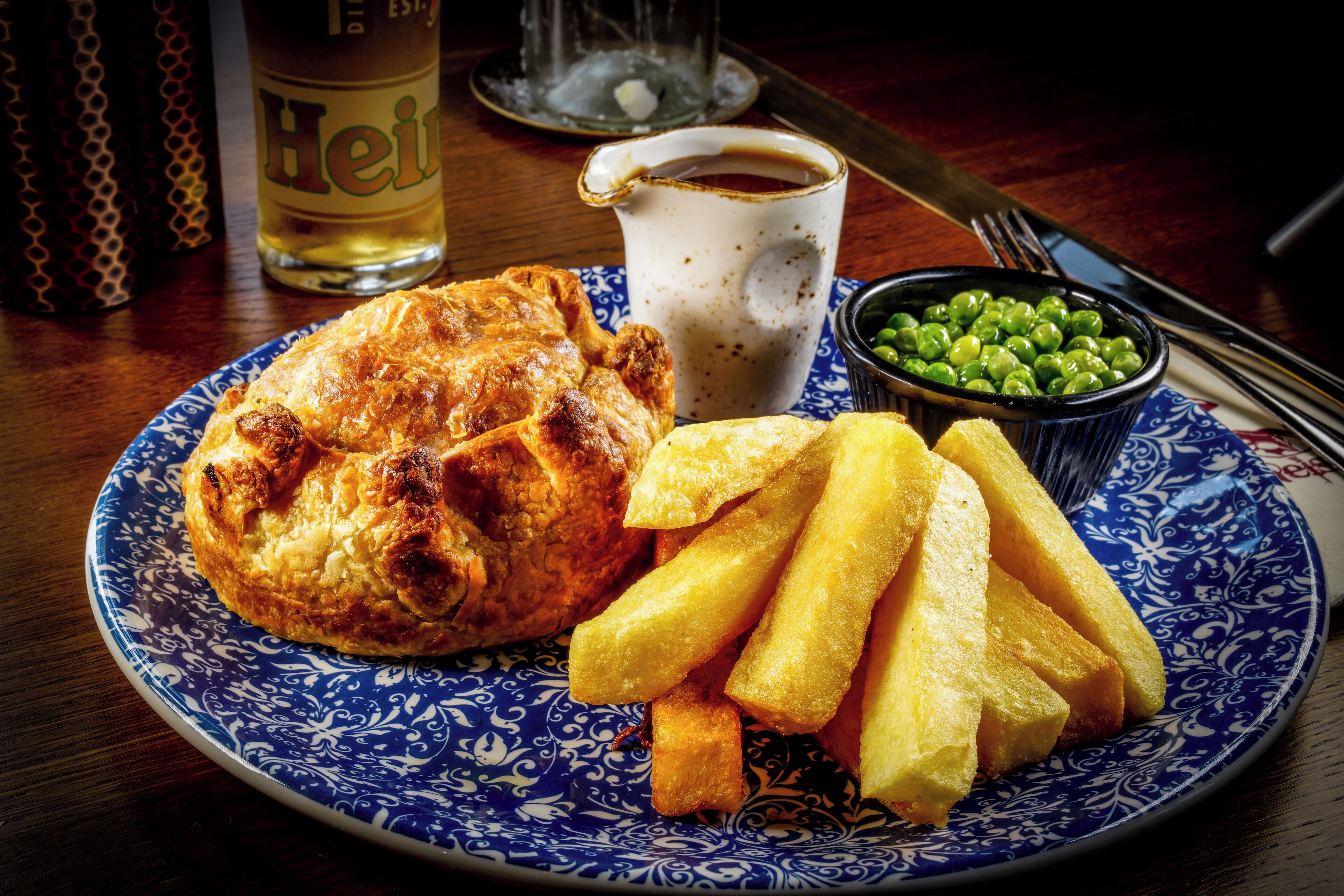 A plate with a baked item, fries, and a small bowl of peas, accompanied by a cup of coffee and a glass of beer on a wooden table.