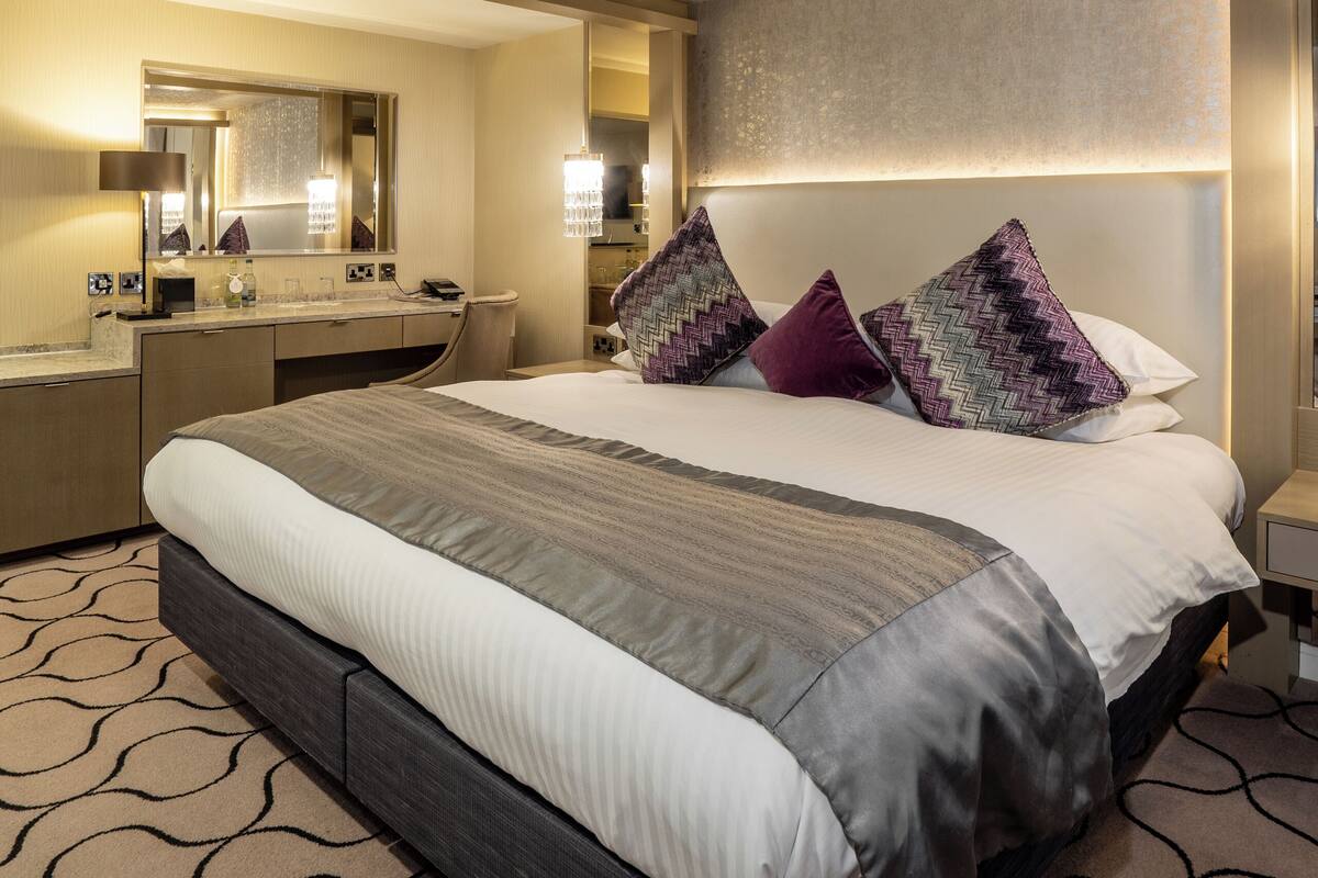 A modern hotel room with a large bed, a bedside table, a mirror, and a headboard with built-in lighting.