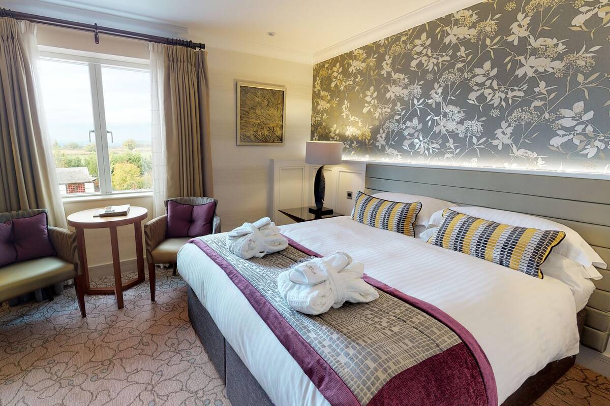 A hotel room with a large bed, two armchairs, a small round table, and a view of the outdoors.