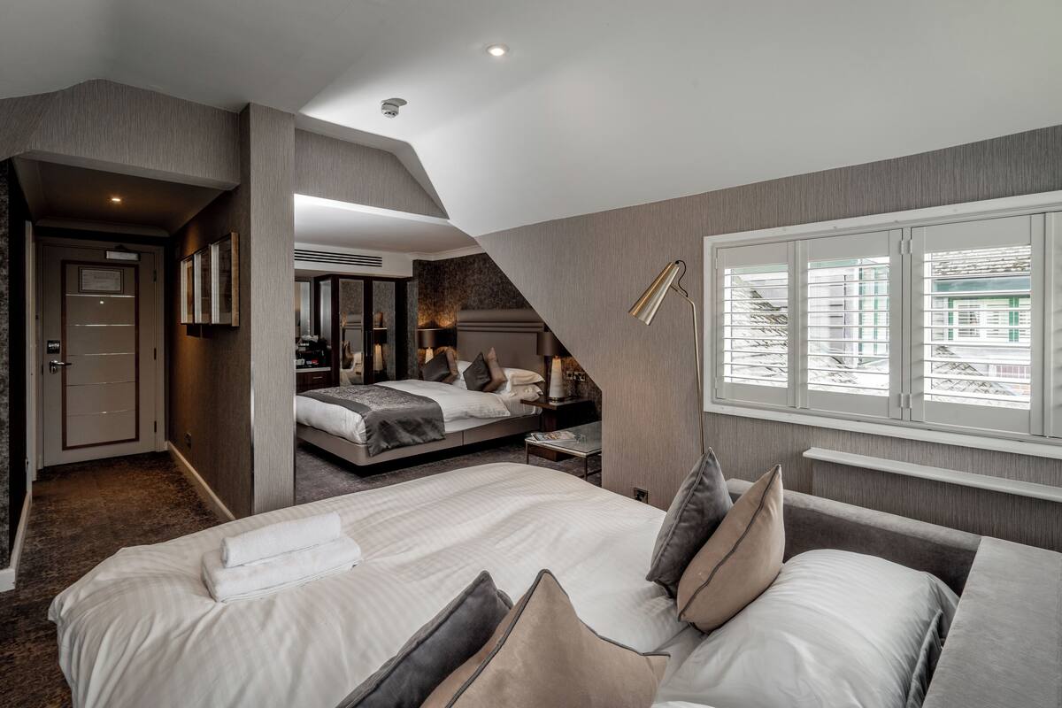 A modern hotel room with two beds, a large window with blinds, a desk, and a mirror.