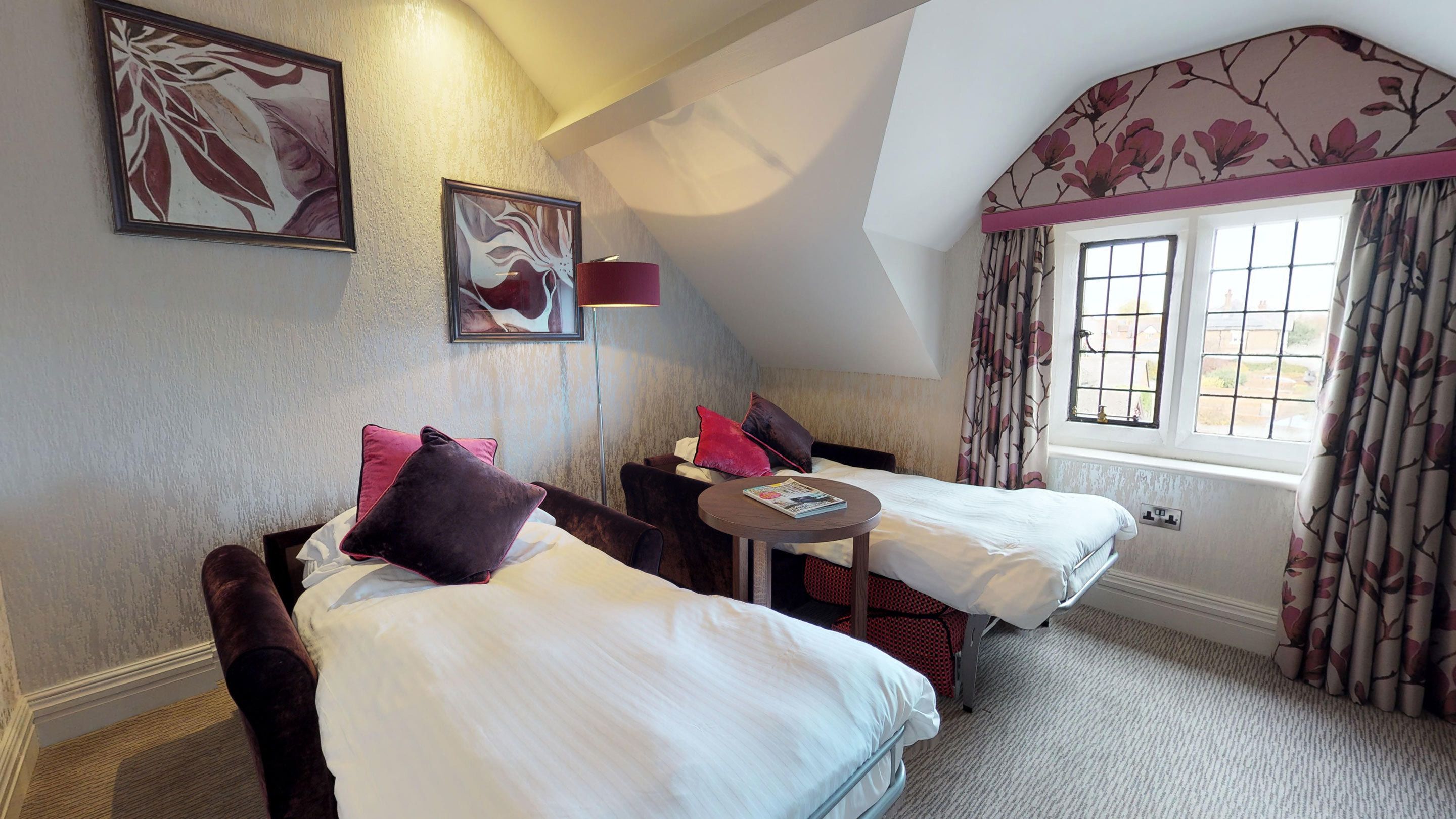 A hotel room with two beds, a small table, and a window with curtains.