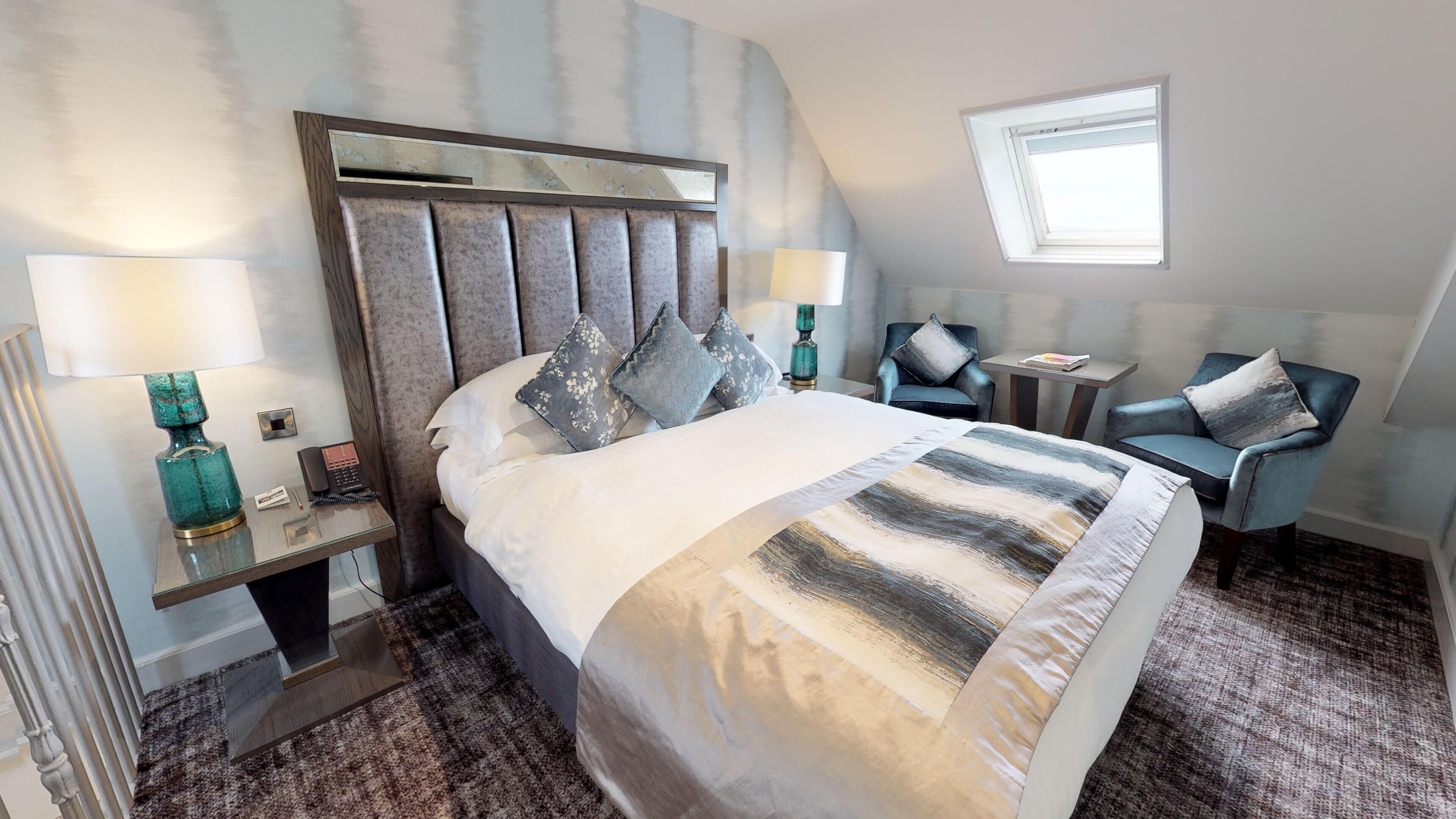 A modern hotel room with a large bed, a sitting area with two chairs, a small table, and a window.