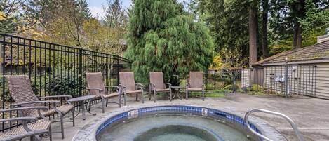 A hot tub surrounded by wicker chairs, with a metal fence and tall trees in the background.