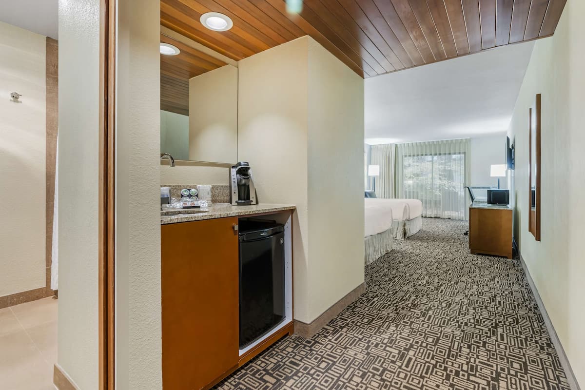 A hotel room with a kitchenette, a bed with a white comforter, a TV, and a window with curtains.