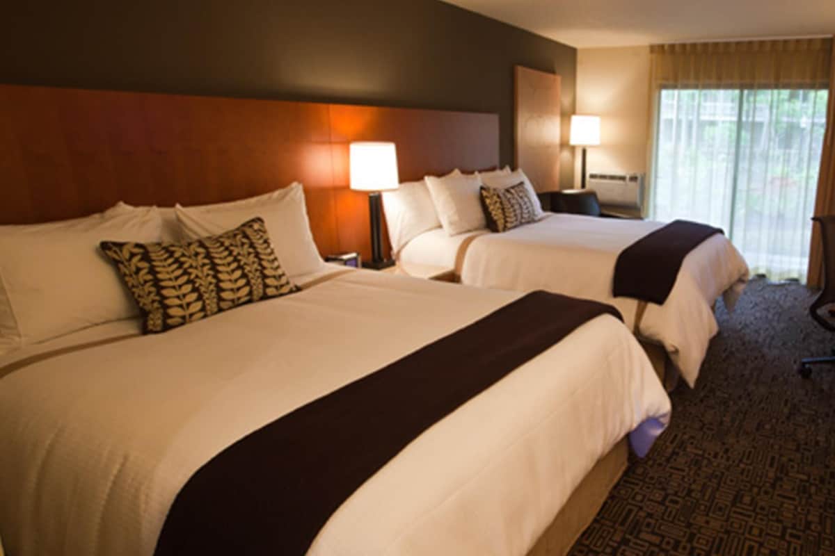 A hotel room with two beds, a headboard, bedside lamps, a carpeted floor, and a window with curtains.