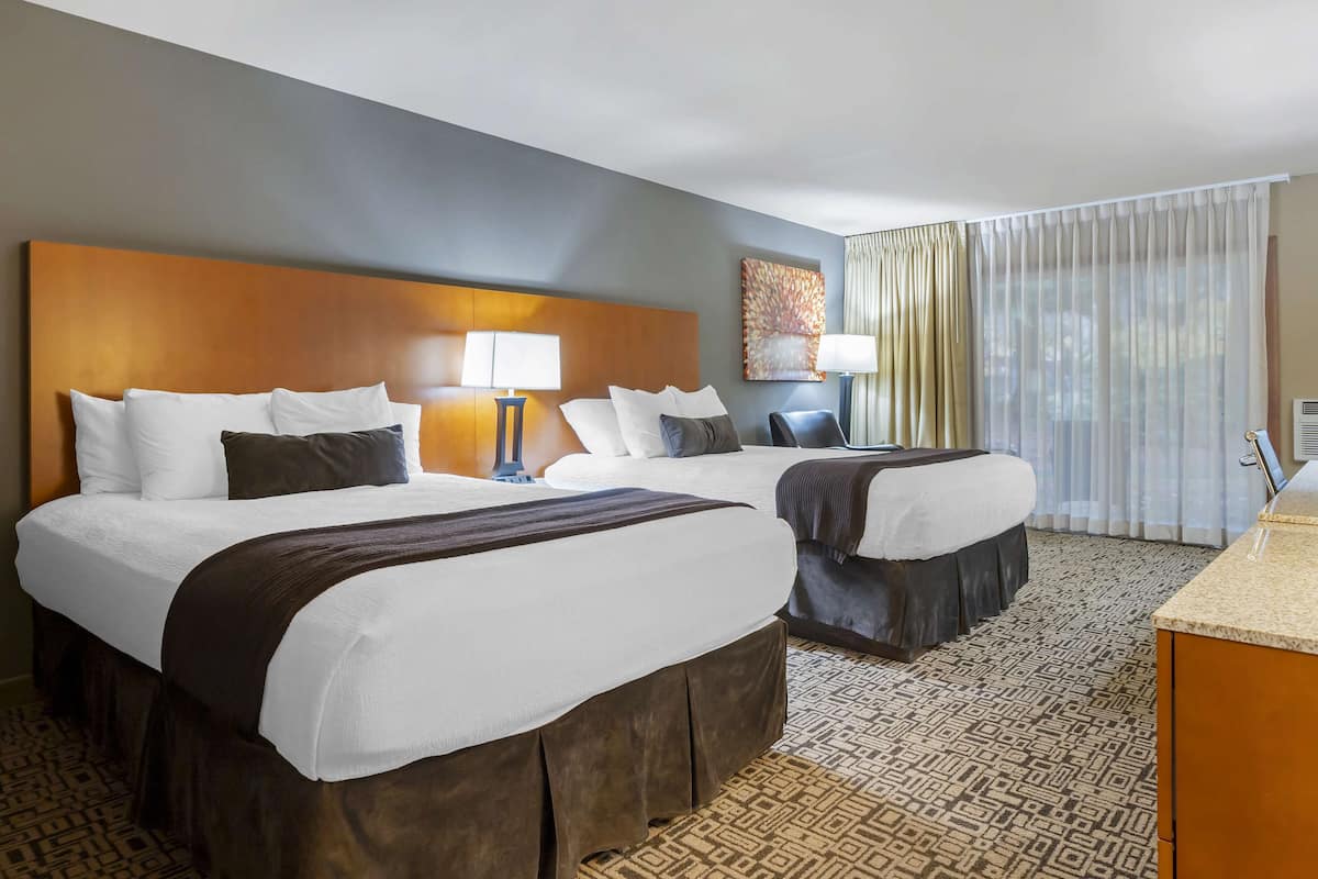 A hotel room with two beds, a headboard, bedside lamps, a painting, a window with curtains, and a patterned carpet.
