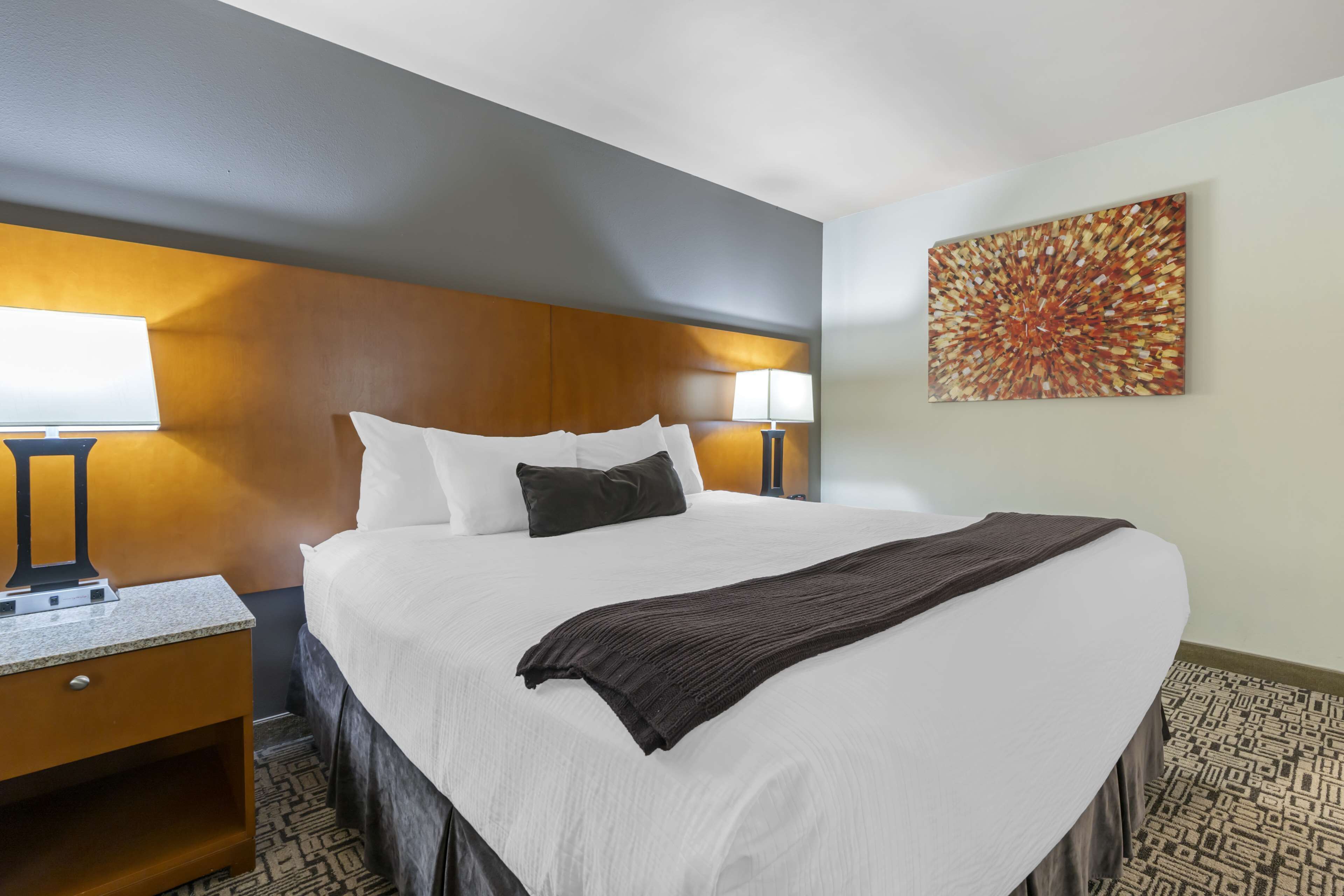 A hotel room with a large bed, two bedside lamps, a headboard, a wall-mounted artwork, and a patterned carpet.