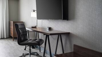 Una habitación de hotel moderna con un televisor de pantalla plana grande montado en la pared, un escritorio de madera, una silla de cuero negro y una mesita de noche pequeña.