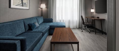 Ein modernes Hotelzimmer mit einer Eckcouch, einem Holztisch, einem Schreibtisch mit Stuhl, einem an der Wand montierten Fernseher und einem großen Fenster mit transparenten Vorhängen.